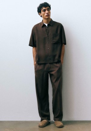 Mladý muž s kučeravými vlasmi, oblečený v tmavohnedej krátkorukávovej košeli na gombíky a zodpovedajúcich nohaviciach, stojaci s rukami v kapse.