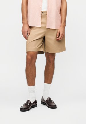 Uomo che indossa pantaloncini beige fino al ginocchio, calze bianche corte, mocassini marrone scuro e una camicia rosa chiaro sbottonata sopra una maglietta bianca.