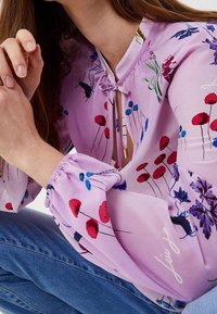 Rosa blus med ett blommönster i röd, blå och grön. Har en framsluten knytning och elastiska ärmar. Bärs med blå jeans.