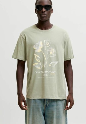 Person iført sorte solbriller, lysegrøn T-shirt med falmet blomsterprint og tekst, kombineret med blå denimjeans, stående mod ensfarvet baggrund.