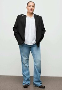 Blazer negro sobre una camisa blanca, combinados con vaqueros acampanados azul claro. El conjunto incluye calzado oscuro y un fondo neutro.