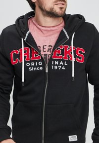 Sudadera con cremallera negra y logotipo bordado en rojo "CREEKS", con rayas de acento blancas y una textura suave. Presenta bolsillos frontales y capucha con cordón.