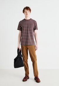 Striped burgundy t-shirt, short sleeves; caramel-colored pants; black quilted bag; brown shoes with tassel detail; plain background.