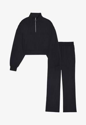 Fekete, rövidített pulóver magas gallérral és ezüst cipzárral, párosítva hasonló anyagból készült, bő szabású nadrággal. Mindkettő puha anyagból készült.