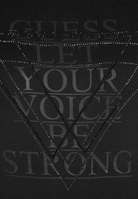 Černé tričko s grafickou potiskem s textem "GUESS LET YOUR VOICE BE STRONG", zdobené lesklými, diamantovými akcenty.