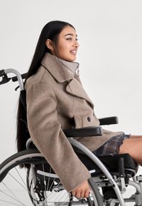 Młoda kobieta w beżowym płaszczu korzystająca z wózka inwalidzkiego, uśmiechnięta i patrząca przed siebie na jednolitym tle.