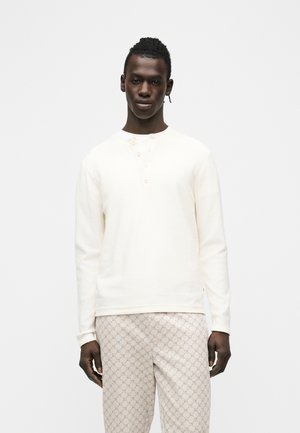 Muž stojaci pred čistým bielym pozadím, oblečený v dlhorukávovej bielej henley košeli a svetlo béžových nohaviciach s vzorom.