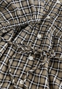 Chemise à carreaux marron et noire avec des accents blancs, dotée d'un col froncé, de boutons sur le devant et d'une texture de tissu douce.