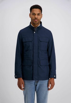 Mann trägt eine dunkelblaue Jacke mit vier aufgesetzten Taschen vorne und hellblaue Jeans, steht vor einem schlichten hellen Hintergrund.