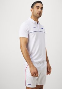 Weißes Poloshirt aus strukturiertem Stoff, mit einem blauen Logo und einem blauen Streifen über der Brust; kombiniert mit weißen Shorts mit roten Akzenten.