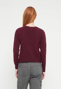 Maglione bordeaux lavorato a maglia con scollo rotondo e maniche lunghe, caratterizzato da un orlo e polsini a costine, abbinato a pantaloni cargo grigi.