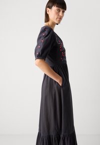 Schwarzes Kleid mit Puffärmeln und floraler Stickerei in Rot und Blau, geraffter Taille und Seitentaschen. Weicher Stoff mit einem gestuften Saum.