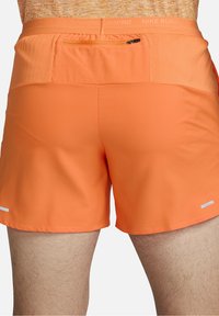 Oranssit juoksushortsit, joissa on teksturoitu vyötärö, vetoketjullinen tasku takana ja heijastavat yksityiskohdat helmassa. Sujuva kangaspinta.