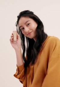 Flacon de fond de teint avec un design en verre transparent, liquide doré pâle à l'intérieur, tenu par une personne portant un haut oversized de couleur moutarde devant un fond clair.