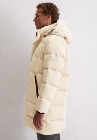 Roomwitte donsjas met capuchon, voorzien van verticale doorgestikte secties, zwarte ritszakken en een subtiel logo op de mouw.