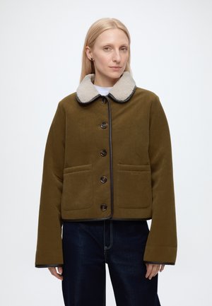 Femme portant une veste kaki boutonnée avec un col en polaire blanc et un jean bleu foncé sur un fond blanc uni.