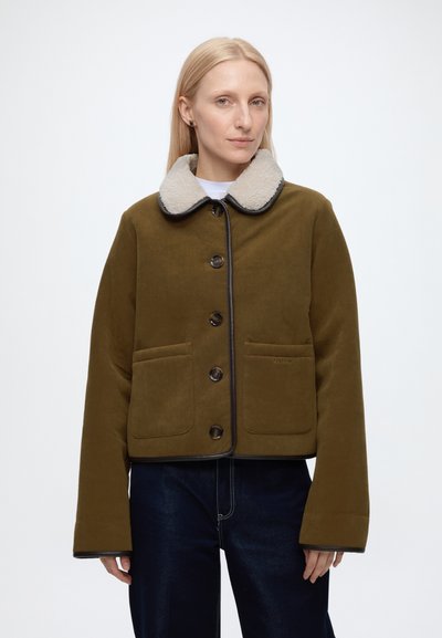 Femme portant une veste kaki boutonnée avec un col en polaire blanc et un jean bleu foncé sur un fond blanc uni.