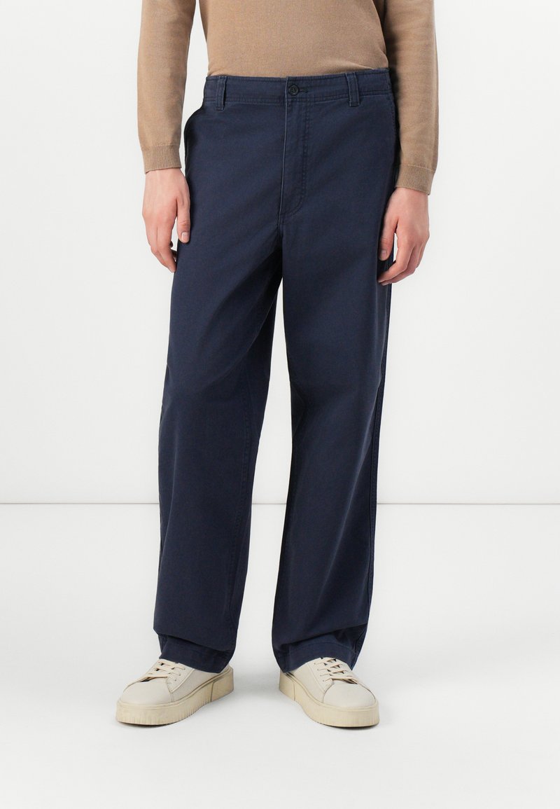 Navy blue chinos with a straight-leg fit, featuring a front button closure, side pockets, and soft cotton fabric. Paired with cream sneakers.