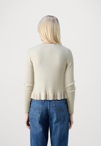 Kremowy sweter z prążkowanego materiału, z długimi rękawami i falbaną u dołu, zestawiony z wysokim stanem niebieskich dżinsów. Widok od tyłu, gładka tekstura.