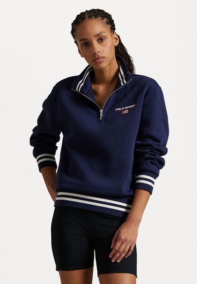 Námořnická modrá mikina se zipem a bílými pruhovanými manžetami a lemem. Obsahuje logo "POLO SPORT" s detailem americké vlajky. Měkká textilní textura.