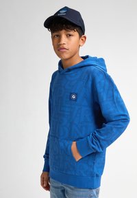 Modrá mikina s kapucí, která má texturovaný grafický potisk s různými písmeny, přední kapsu a žebrované manžety. V kombinaci s tmavou čepicí.