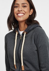Grå zip-up hoodie med en kontrasterande stickad huva, krämfärgade repdragband och läderdetaljer på snörena, med en mjuk textur.