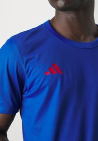 T-shirt sportivo blu realizzato in materiale liscio e traspirante, con scollatura rotonda e un prominente logo rosso a tre strisce sul petto.
