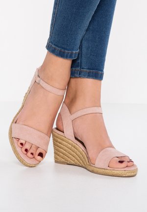 Pieds dans des sandales compensées en daim rose avec des lanières aux chevilles et des ongles de pieds peints en rouge foncé, portant un jean denim court bleu.