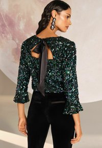 Pantaloni in velluto nero abbinati a un top in paillettes verdi con schiena a oblò, legato con un nastro di raso e maniche svasate.