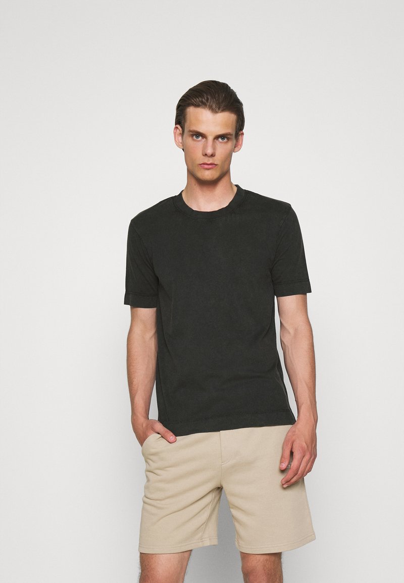 Zwarte korte mouwen t-shirt gemaakt van katoen, met een ronde hals en een ontspannen pasvorm, gecombineerd met beige shorts, tegen een lichte achtergrond.