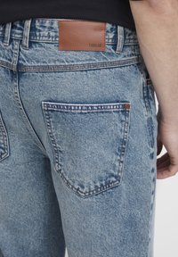 Hämarad sinised teksapüksid koos beežikarva nahast sildiga, millel on tavalised tagataskud ja õmblustega detailid õmbluste ääres.