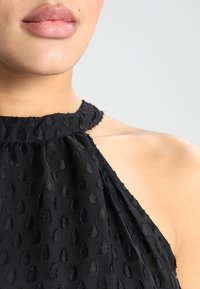 Musta halterneck-toppi, joka on valmistettu tiheästä kankaita, jossa on kohoavia pisaramaisia kuvioita. Sujuva kaula-aukko ja laskostettu muotoilu korostavat sen muotoa.