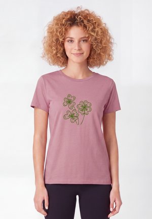 Vrouw met krullend haar die een mauve t-shirt draagt met een groen ontwerp van een drie- of vierbladige klaver, gecombineerd met een zwarte broek tegen een lichte achtergrond.