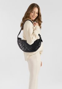 Borsa a mano in pelle nera con un motivo trapuntato, forma a mezzaluna e una tracolla intrecciata, esposta su uno sfondo neutro.