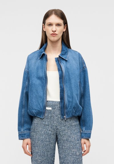 Jeune femme portant une veste zippée en jean bleu sur un haut blanc et un pantalon texturé taille haute, debout devant un fond uni.