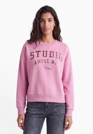 Femme aux longs cheveux foncés portant un sweat-shirt rose avec un texte métallique et un jean gris foncé, debout sur un fond blanc.