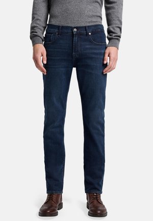 Jeans en denim bleu foncé à coupe slim, avec des poches standards, une fermeture à bouton en métal et un léger effet vieilli. Accompagnés de chaussures en cuir marron.