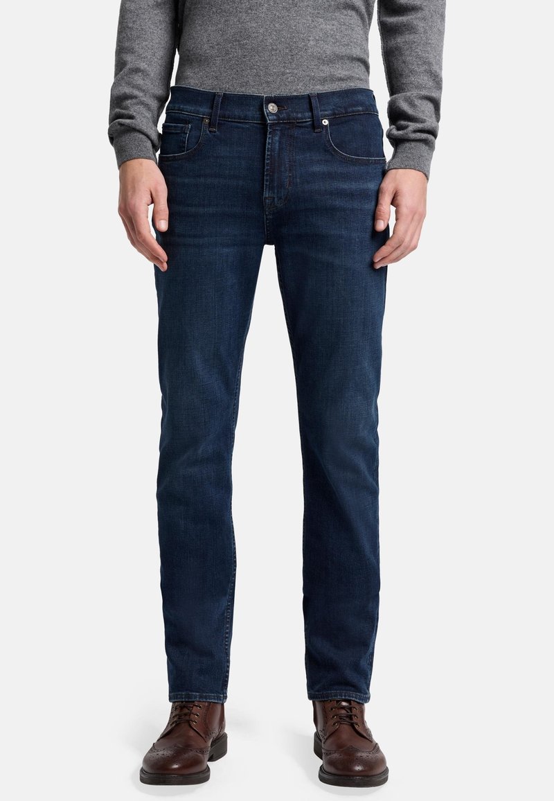 Dunkelblaue Denim-Jeans im Slim-Fit, mit Standardtaschen, Metallknopfverschluss und minimalen Abnutzungserscheinungen. Kombiniert mit braunen Lederschuhen.