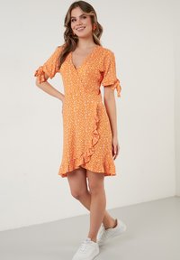 Oranjekleurige bloemenjurk met een V-hals, gerimpelde zoom en korte mouwen die bij de manchetten zijn gestrikt. Gecombineerd met witte sneakers op een neutrale achtergrond.