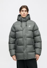 Jaqueta puffer cinza com capuz destacável, bolso no peito com fecho éclair e detalhes com cordão. Feita de um material liso e leve.