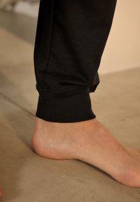 Marginile pantalonilor sport din bumbac negru se odihnesc lângă un picior gol pe o suprafață de beton, având o textură netedă și detaliu simplu de cusătură.