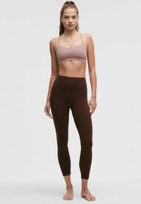 Bruine high-waisted leggings gecombineerd met een lichtroze sportbeha met dunne bandjes. De materialen lijken rekbaar en glad.