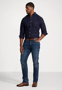 Temno modra srajca na gumbe, modre kavbojke, rjav usnjen pas in svetlo rjavi mokasini. Srajca ima ovratnik in logotip na prsih.