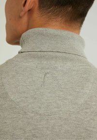 Pull gris en maille avec un col montant côtelé, présentant un discret logo cousu à la nuque et une texture lisse et uniforme sur toute la surface.
