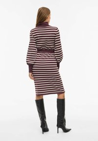 Kootud kleit kõrge kaelusega, millel on horisontaalsed marooni ja kreemi triibud, liibuv vöökoht ja balloonvarrukad, kombineeritud mustade põlvini ulatuvate saapadega.