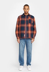 Camisa de cuadros en azul marino y naranja quemado, diseño de botones con un bolsillo en el pecho. Llevada sobre una camisa gris con pantalones de denim azul y zapatillas blancas.