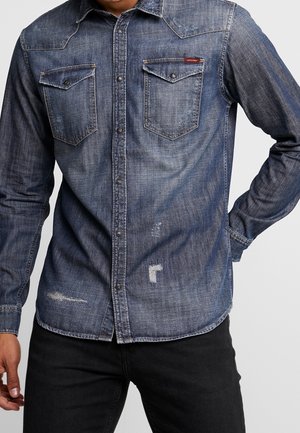 Man wearing a faded blue denim button-up shirt with two chest flap pockets and distressed details, paired with black pants.