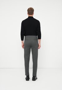 Bărbat îmbrăcat în pantaloni gri slim-fit, bluză neagră cu mânecă lungă și pantofi negri, stând cu fața spre un zid alb, cu mâinile pe lângă corp.