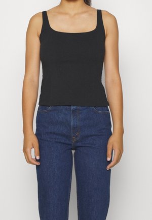 Kobieta w dopasowanym czarnym topie bez rękawów z kwadratowym dekoltem i niebieskich dżinsach z wysokim stanem, stojąca na jednolitym tle.