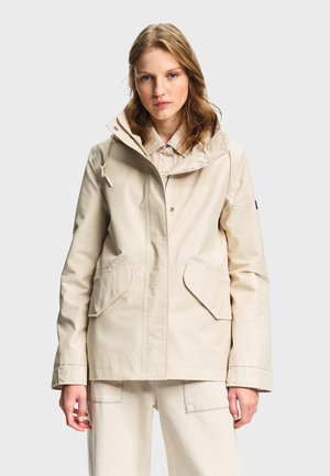 Jeune femme portant une veste à capuche beige avec des poches à rabat sur le devant et un pantalon clair assorti, sur un fond uni.